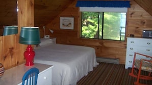 Ein Schlafzimmer in einer Holzhütte mit Bett, Kommode, Stuhl, Lampe und einem Fenster mit Blick auf Bäume.