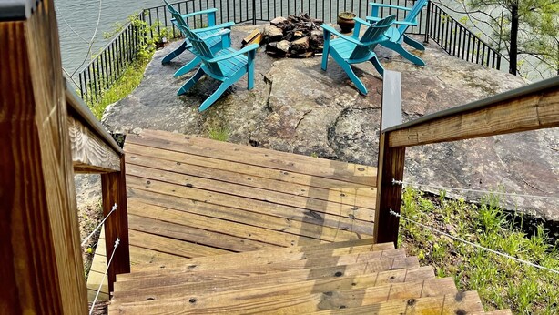 Una terraza de madera con sillas Adirondack azules, un hogar para fuego y vistas a un lago con barcos y casas al fondo.