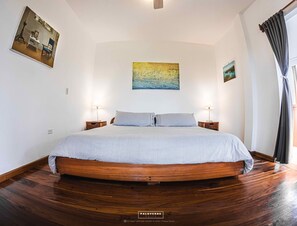A bedroom with a large bed, two bedside tables with lamps, a wooden floor, and artwork on the walls.