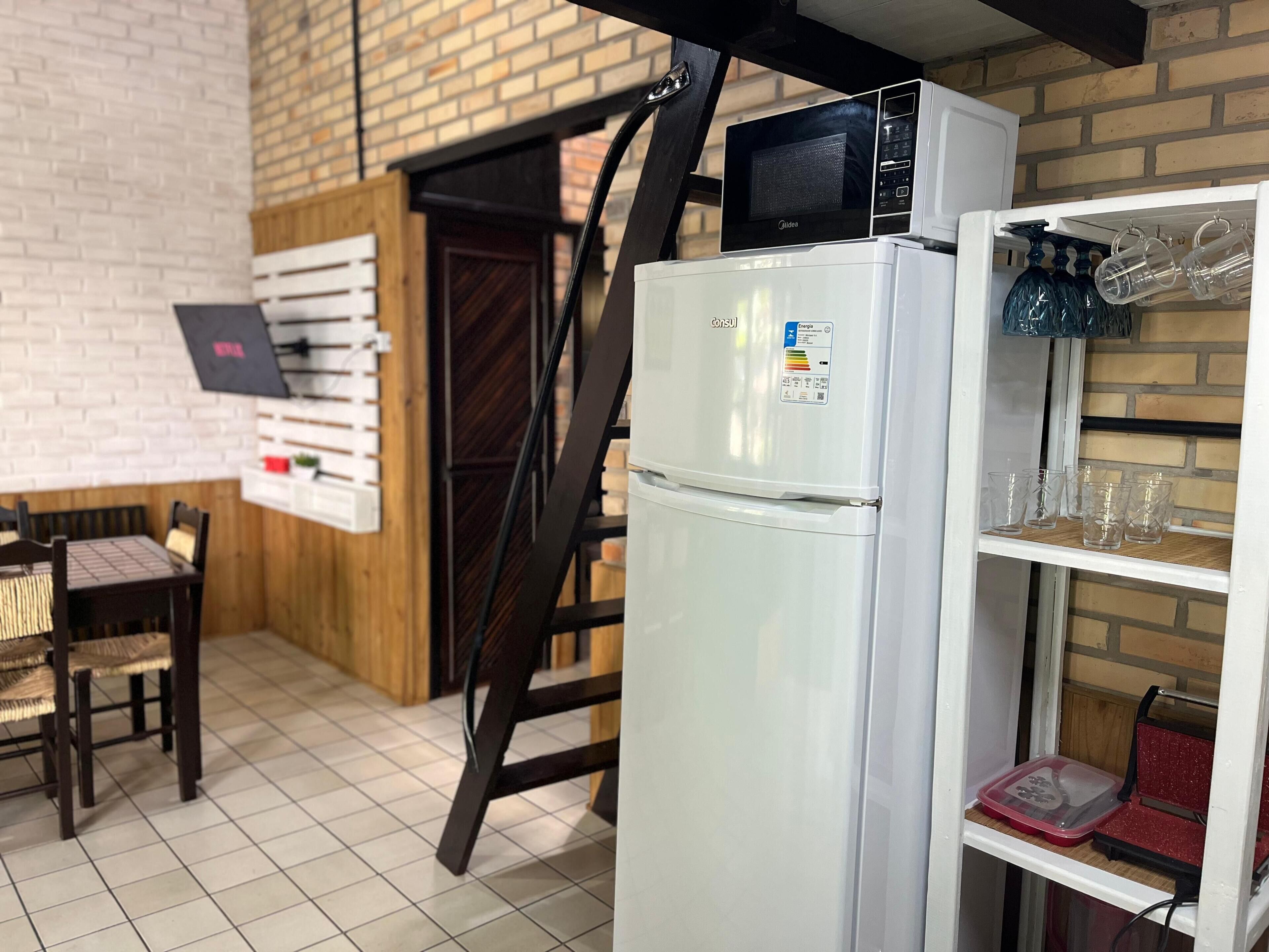 A kitchen with a refrigerator, microwave, and a shelf with glasses and a red container.