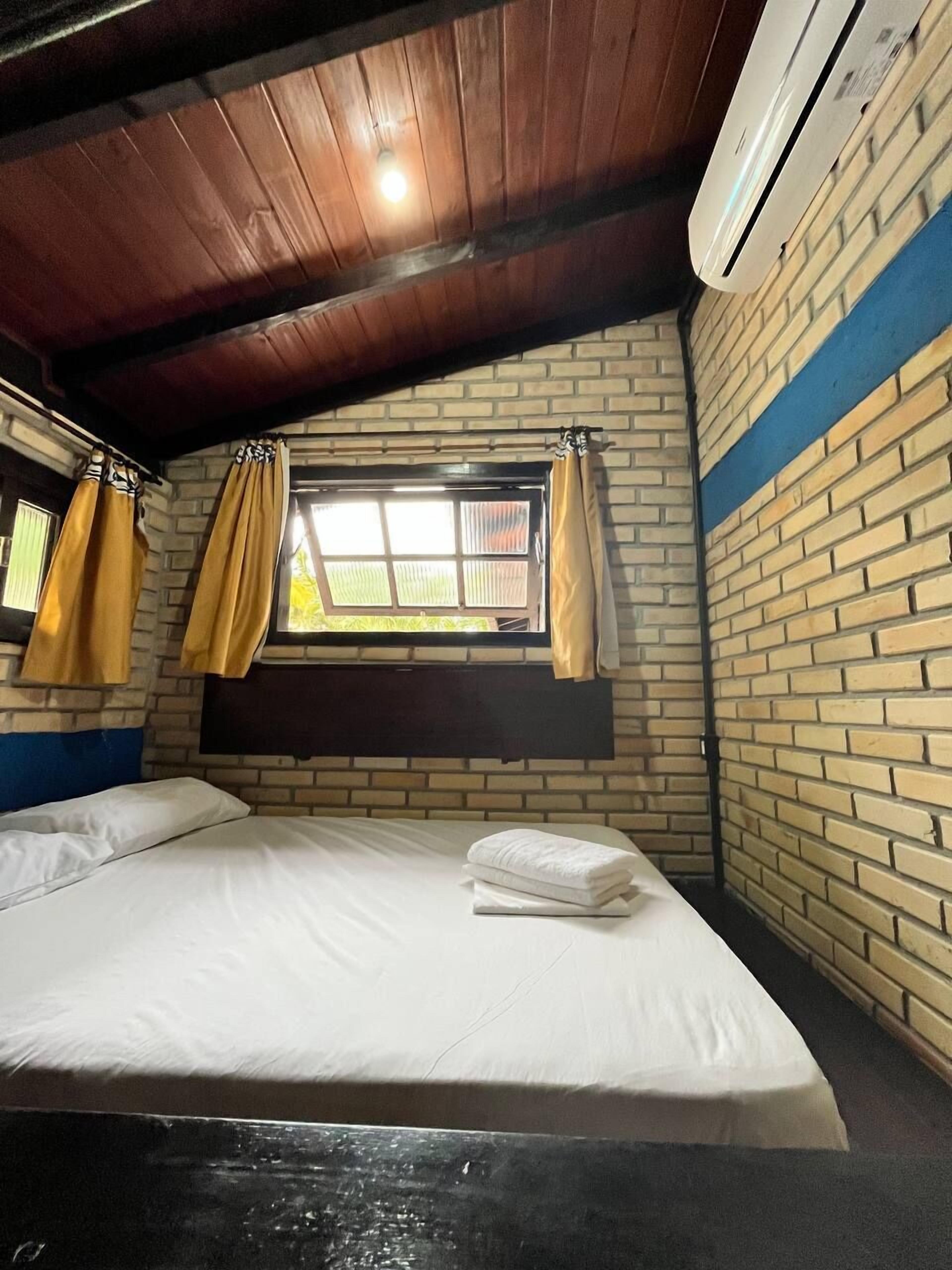 Una cama individual en una habitación con pared de ladrillo, ventana con cortinas amarillas y un equipo de aire acondicionado.
