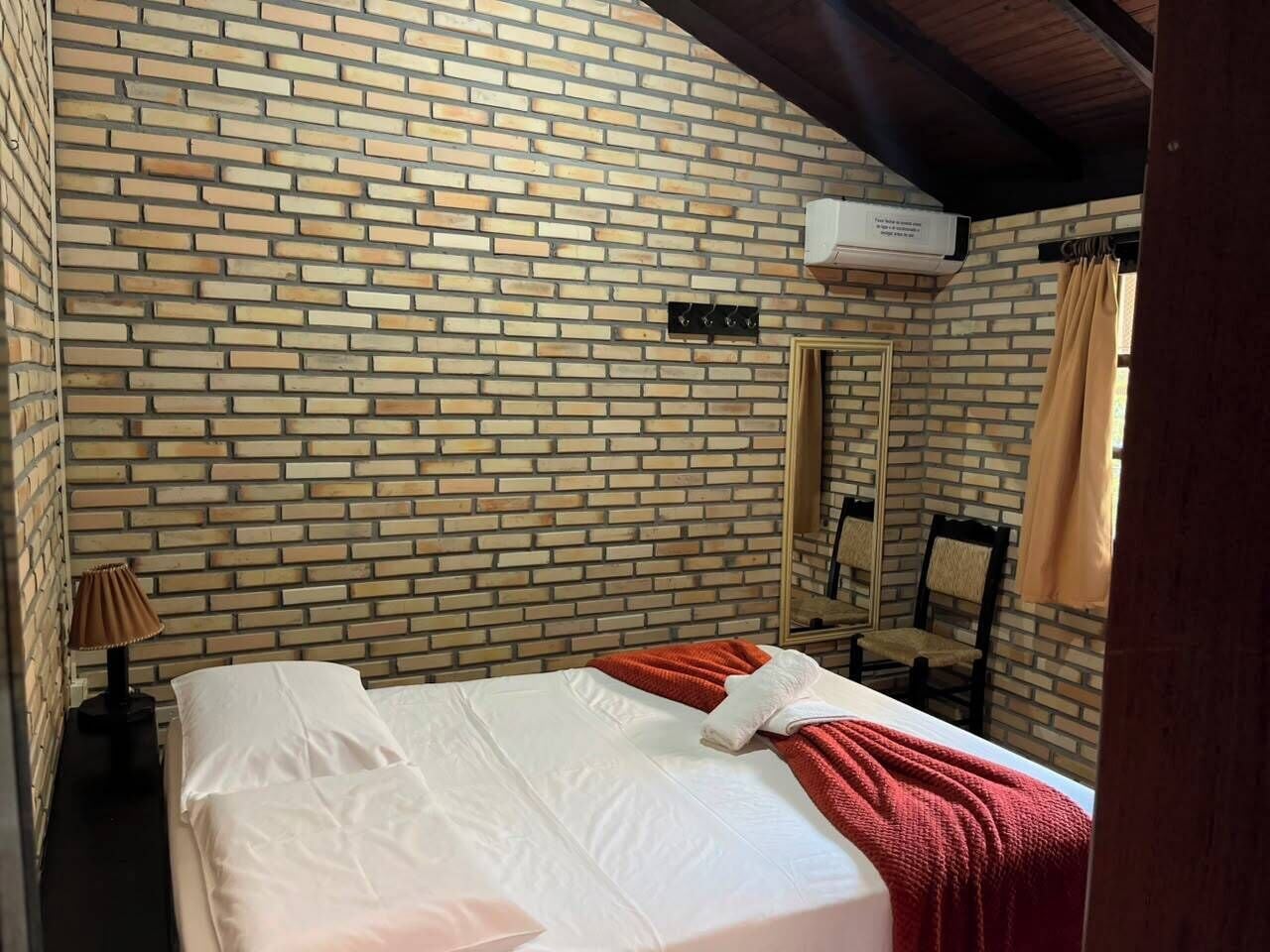 Una habitación de hotel con pared de ladrillo, una cama con ropa blanca, una manta roja, una lámpara de noche y un espejo.