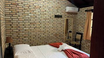 Una habitación de hotel con pared de ladrillo, una cama con ropa blanca, una manta roja, una lámpara de noche y un espejo.