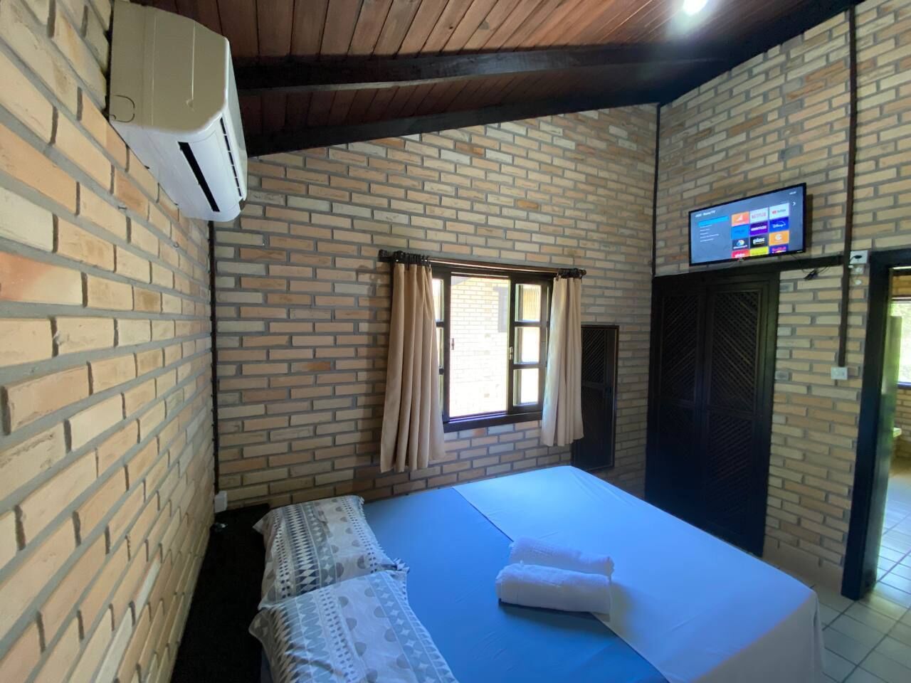 A room with a bed, a window with curtains, a television on the wall, and a brick wall.