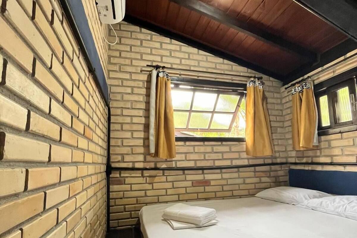 Ein Zimmer mit Backsteinwand, einem Bett mit weißer Bettwäsche, einem Fenster mit Vorhängen und einer Klimaanlage.