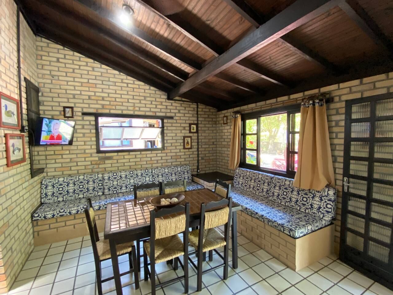 A dining area with a brick wall, wooden ceiling, and a television mounted on the wall.