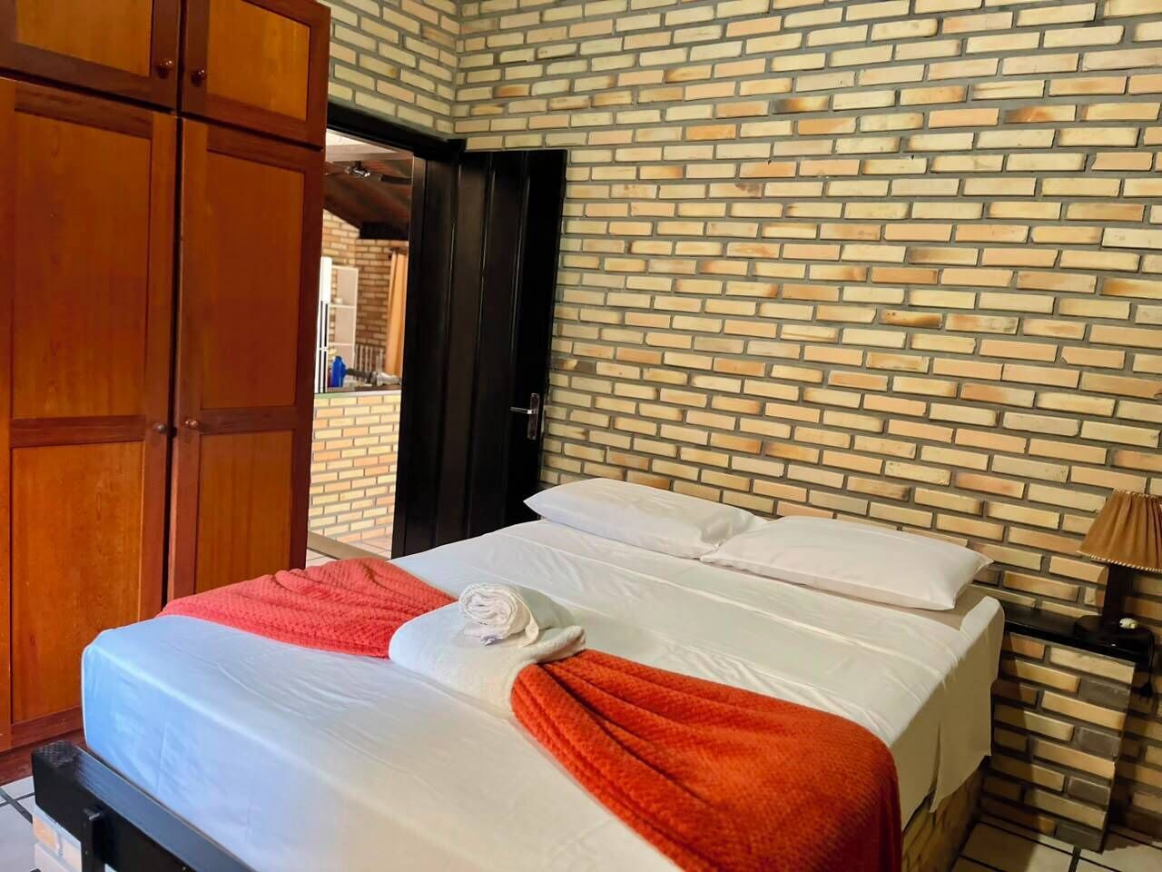 A hotel room with a brick-patterned wall, a bed with white linens, a red blanket, and a wooden wardrobe.