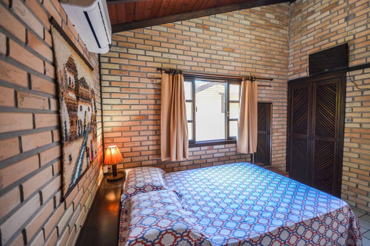 Ein Schlafzimmer mit Ziegelwand, einem Bett mit gemusterten Betttextilien, einem Fenster mit Vorhängen und einer Wand-Klimaanlage.