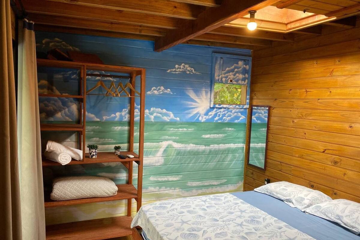 Ein Zimmer mit einem Bett, einer Holzablage mit Handtüchern und einem Wandbild einer Strandlandschaft.