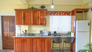 A kitchen with wooden cabinets, a granite countertop, a refrigerator, a microwave, a coffee maker, and a window with blinds. - Eden Villas (Corinth)