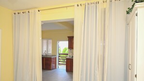 A bedroom with a bed, curtains, and a view into a kitchen and dining area. - Eden Villas (Corinth)