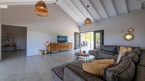 A spacious living room with a large sectional sofa, a wooden cabinet, and a television. There are two hanging pendant lights and a large window with blinds.