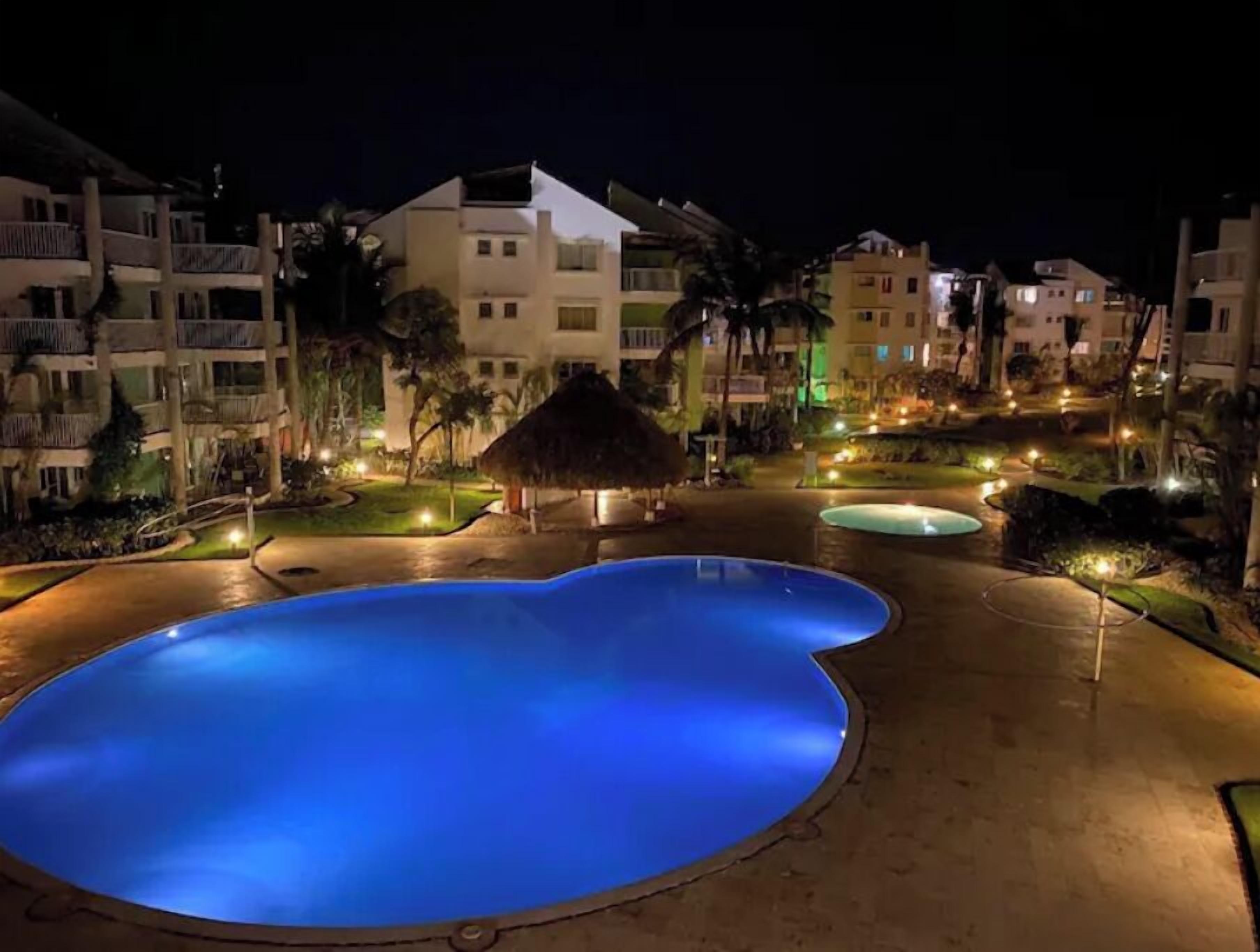 A nighttime view of a resort with a large illuminated swimming pool, palm trees, and multi-story buildings with balconies.