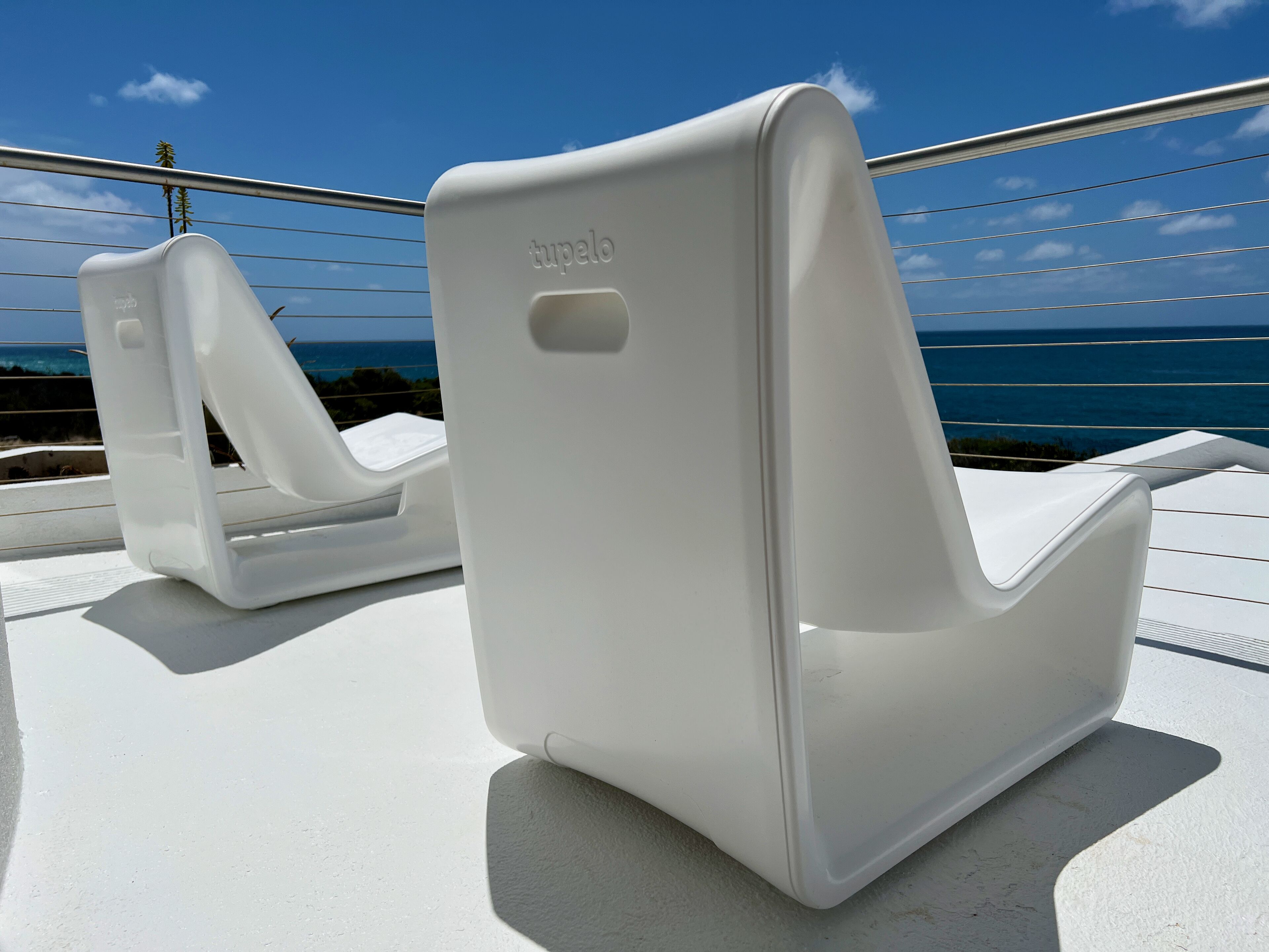 Zwei weiße Lounge-Stühle mit dem Markennamen 'tupe' auf der Rückenlehne, auf einem Balkon mit Blick aufs Meer.