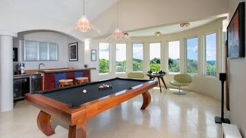 A pool table in a spacious room with large windows offering a view of greenery.
