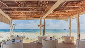 Eine Strandlounge mit runden Tischen und Stühlen, einer Holzbeschichtung und Blick aufs Meer.