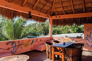 A thatched-roof outdoor seating area with a mural, wooden furniture, and a view of palm trees.