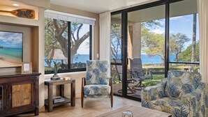 A room with a large window offering a view of the ocean and trees, a wooden coffee table, a chair with a tropical print, and a small table with a lamp.