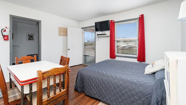 A bedroom with a bed, a TV mounted on the wall, a window with red curtains, and a door leading to another room.
