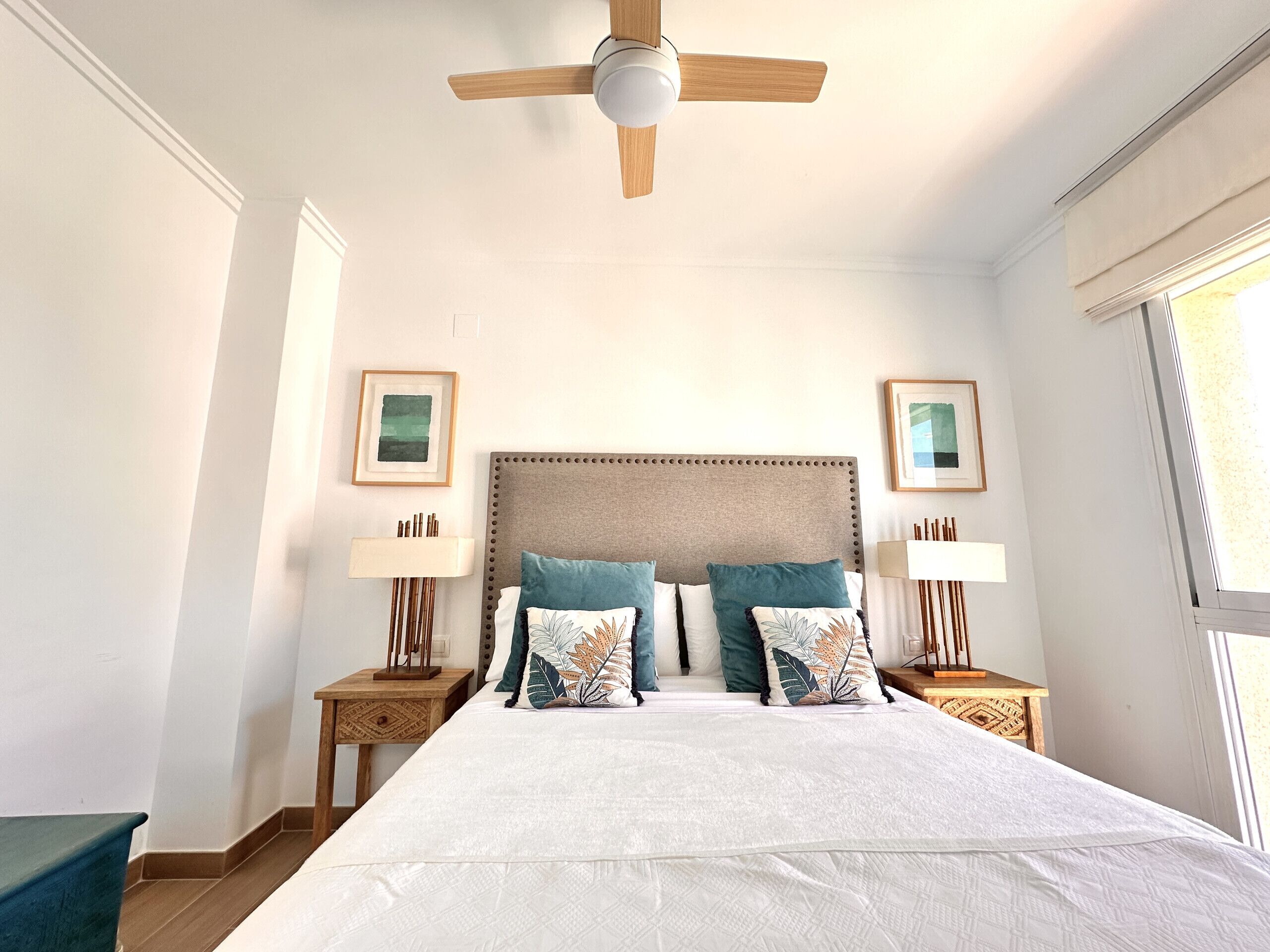 Une chambre avec un grand lit, un ventilateur de plafond en bois, deux tables de chevet avec des lampes et des œuvres d’art encadrées accrochées aux murs.