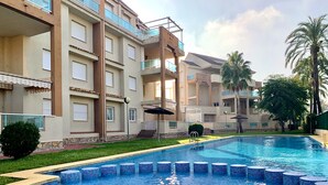 A swimming pool with blue mosaic tiles, surrounded by a building with balconies and a palm tree. - Anacasa Les Marines Puerta del Palmar AP2207 (Denia)