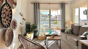 A modern living room with a sofa, a chair, a table, and a balcony with a view of buildings. - Anacasa Les Marines Puerta del Palmar AP2207 (Denia)