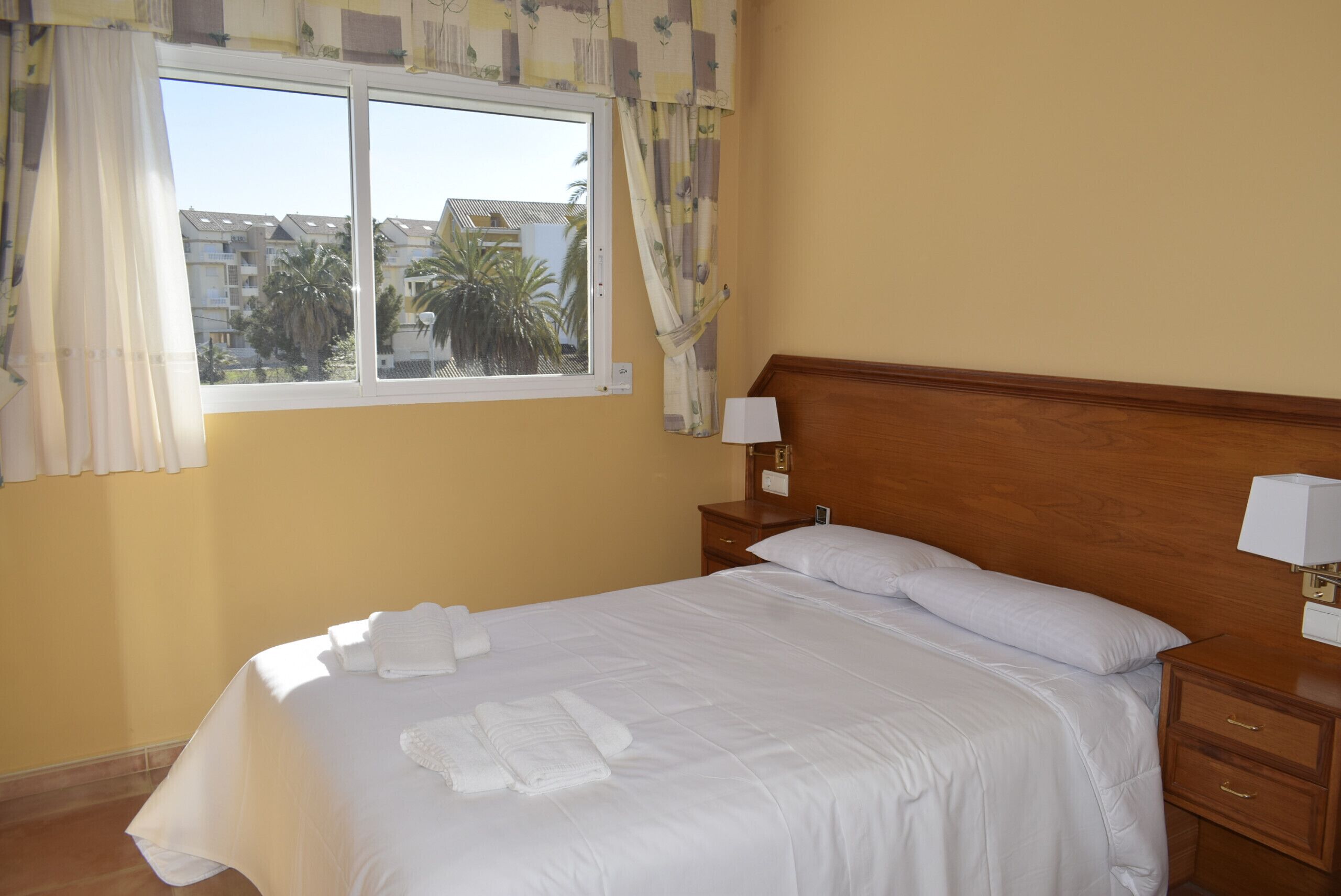 Una cama bien hecha con ropa de cama y toallas blancas, un cabecero de madera, una mesita de noche con lámpara y una ventana con cortinas.