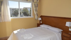 Una cama bien hecha con ropa de cama y toallas blancas, un cabecero de madera, una mesita de noche con lámpara y una ventana con cortinas.