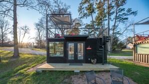 A compact cabin with a deck, a small outdoor seating area, and a view of the surrounding trees. - Combo Plate: 2 mins to Lake Conroe! Heated Pool, Stocked Ponds, Cute Pygmy Goats (Willis)