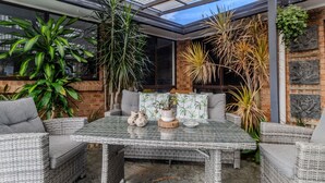 Eine überdachte Terrasse mit Korbmöbeln, einem Glastisch und verschiedenen topfgebundenen Pflanzen.