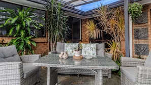 Un patio cubierto con mobiliario de mimbre, una mesa con cubierta de vidrio y diversas plantas en macetas.