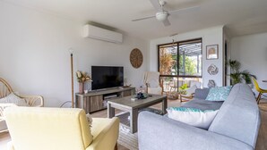 Ein modernes Wohnzimmer mit einer Couch, einem Couchtisch und einem Fernseher. Es gibt einen Deckenventilator, ein großes Fenster mit Blick ins Freie und ein dekoratives Wandbild.