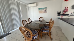 A dining area with a glass table surrounded by wicker chairs with patterned cushions, a painting on the wall, and a fire safety sign. - Avenues Unit 1 (South West Rocks)