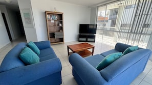 A living room with two blue sofas, a coffee table, a bookshelf, and a TV.