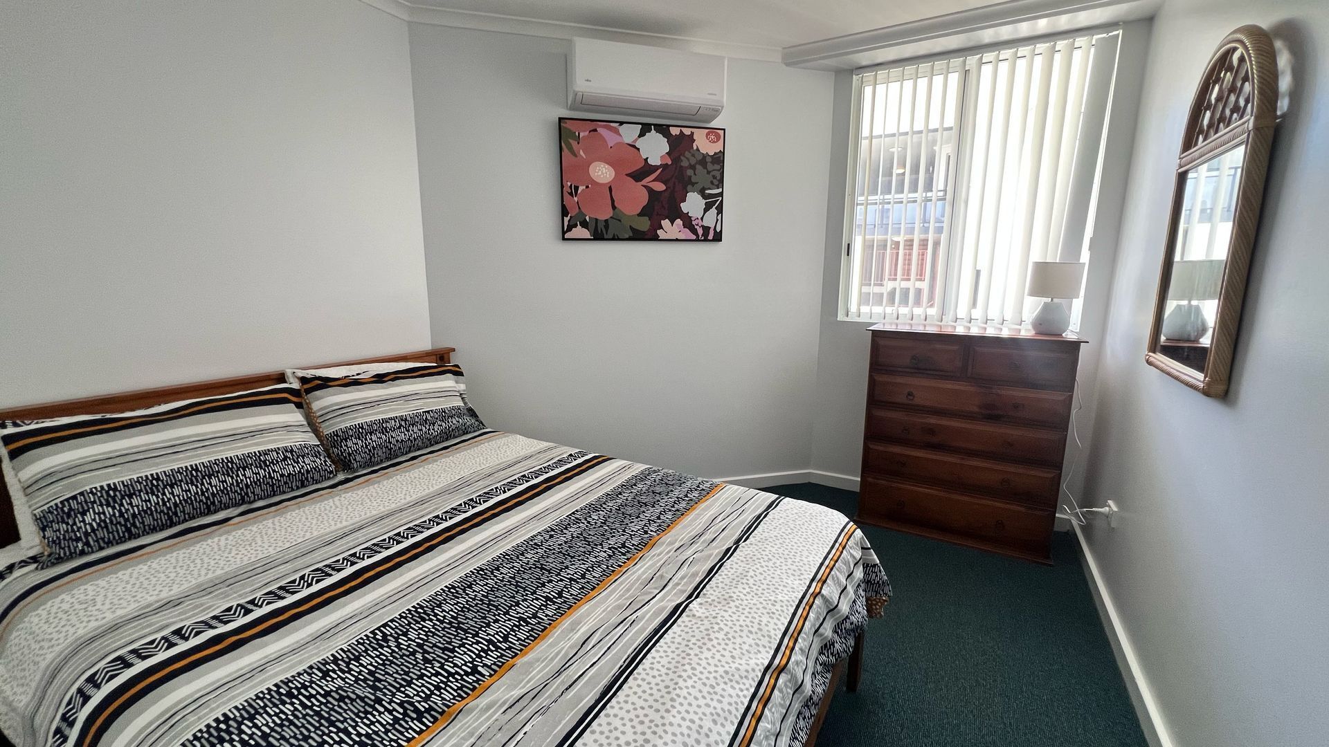 A bedroom with a bed, dresser, window with blinds, and a wall-mounted mirror.