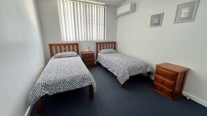 A bedroom with two single beds, a nightstand, a dresser, a window with blinds, and a wall-mounted air conditioner. - Avenues Unit 1 (South West Rocks)