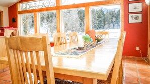 Un comedor con una mesa de madera, sillas y vistas a árboles y montañas a través de amplias ventanas.