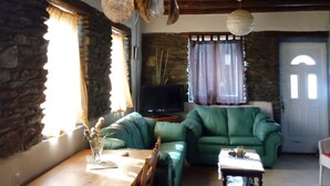 A cozy living room with a stone wall, a green leather sofa, a wooden table, and a hanging light fixture. - Summer Sunrice Andros (Andros)