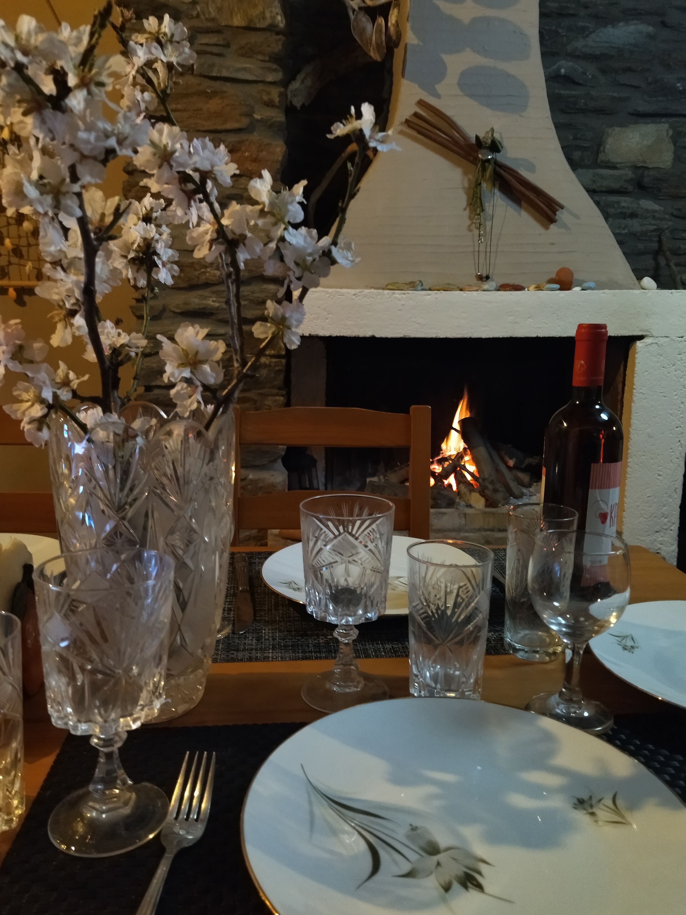 A dining table set with crystal glassware, a vase with white flowers, and a bottle of wine, with a stone wall and fireplace in the background.