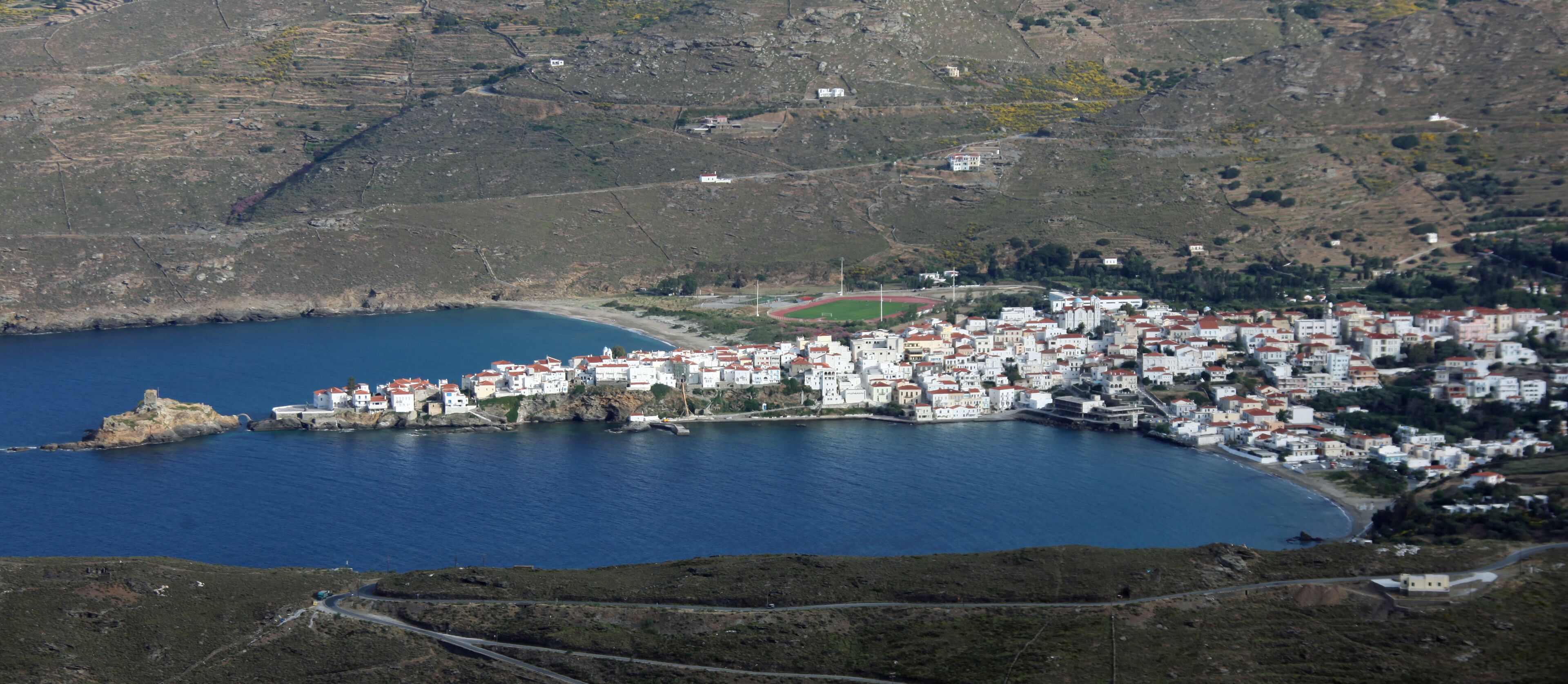 A coastal town with white buildings, a small harbor, and a mountainous backdrop.