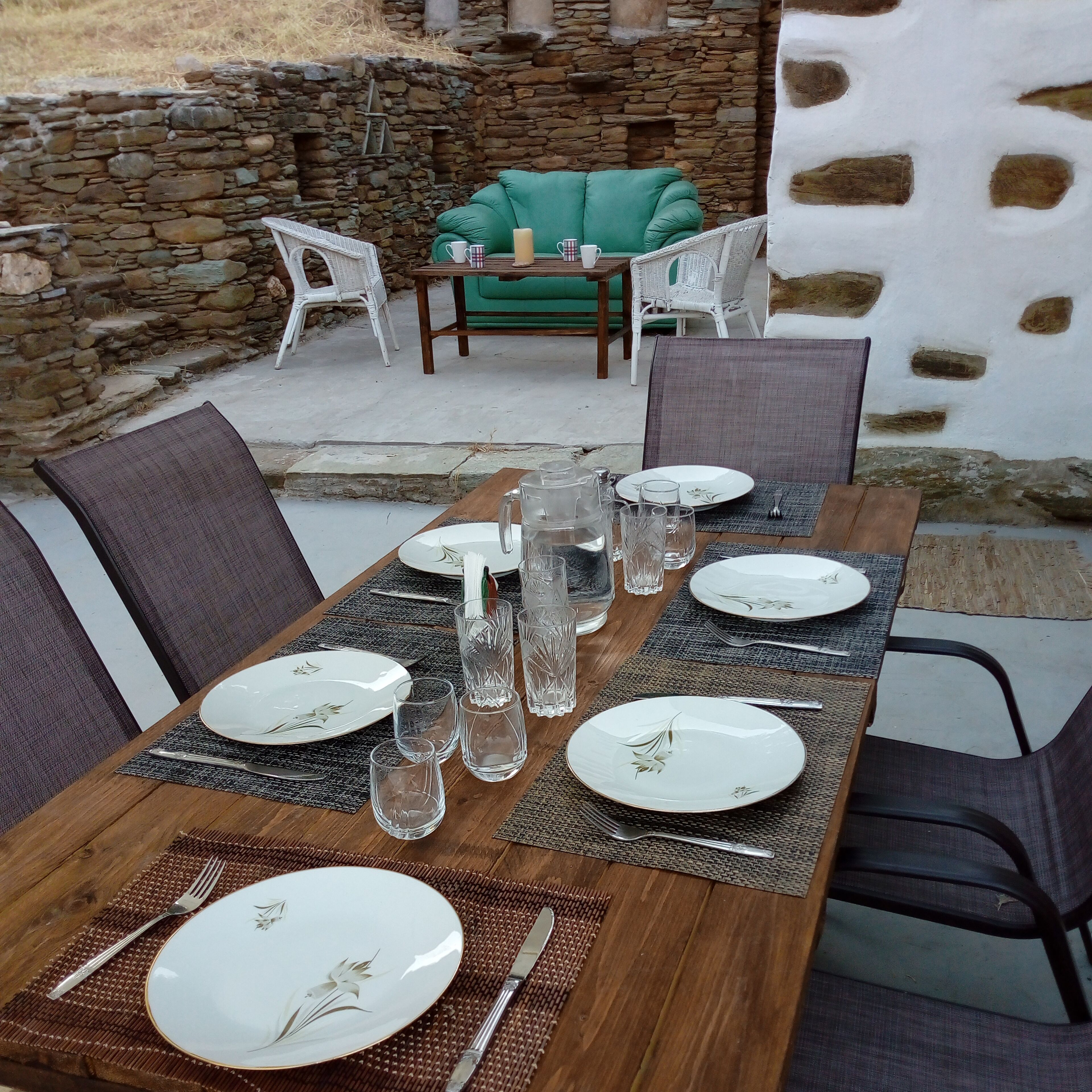 Una mesa de comedor dispuesta con platos, vasos y cubiertos, rodeada de sillas de mimbre y un sofá verde en un espacio al aire libre con paredes de piedra.