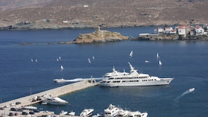 A marina with multiple yachts docked, a pier with parked cars, and a rocky island with a small building in the background. - Summer Sunrice Andros (Andros)