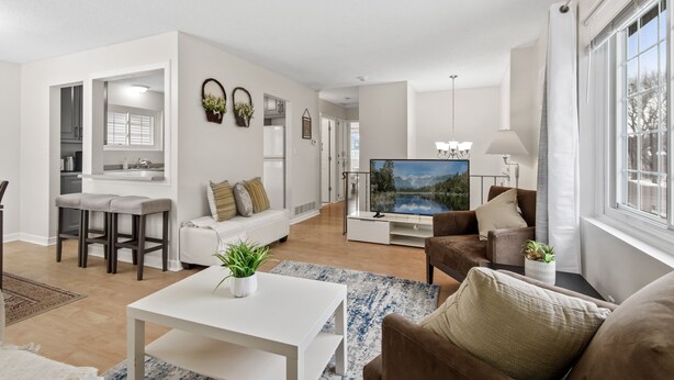 Uma sala de estar moderna com um sofá branco, uma poltrona marrom, um espaço de jantar com uma mesa branca e uma TV exibindo uma paisagem cênica.