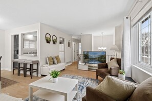 Ein modernes Wohnzimmer mit einem weißen Sofa, einem braunen Sessel, einem Essbereich mit einem weißen Tisch und einem Fernseher, der eine idyllische Landschaft zeigt.