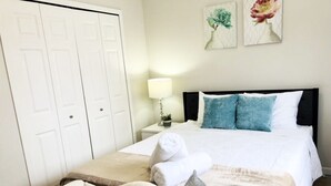 Quarto bem arrumado com cama, almofadas, mesa de cabeceira com abajur e toalha dobrada.