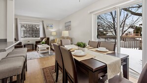Uma sala de jantar com uma mesa grande, cadeiras e um lustre. Há uma janela com cortinas, uma planta na mesa e vista para o exterior.