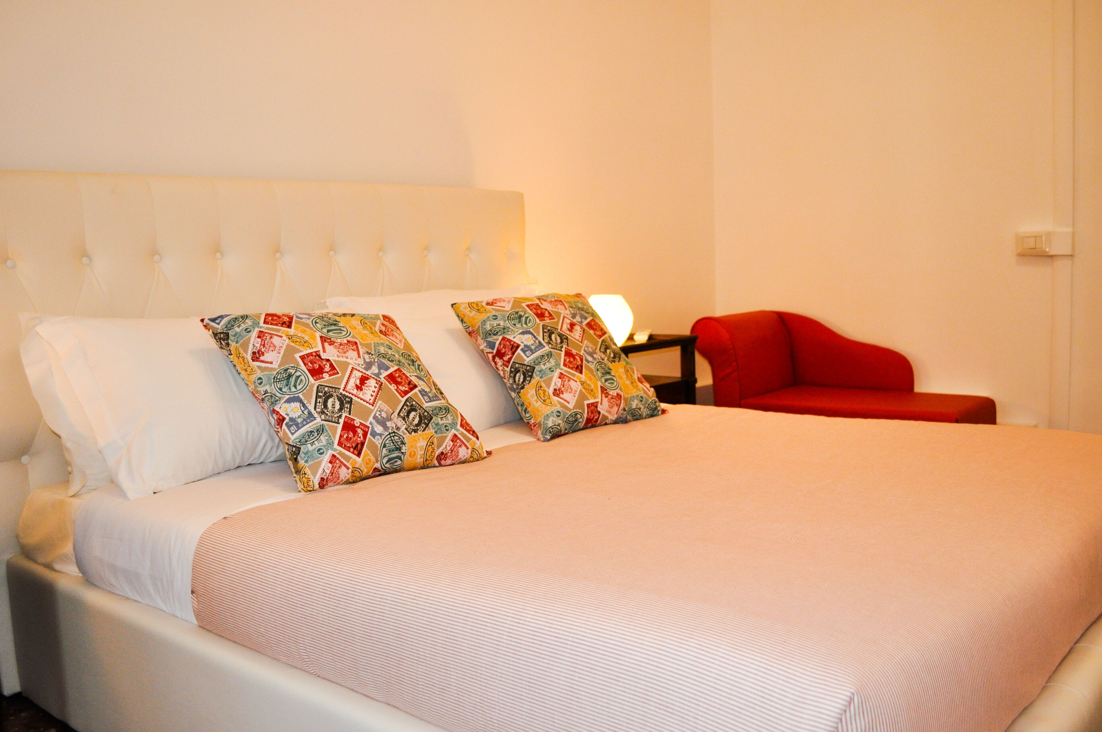 Un lit avec une literie blanche et des coussins aux motifs colorés, une chaise rouge et une table de chevet avec une lampe.