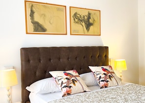 Une chambre à coucher avec une tête de lit capitonnée, des coussins à motifs floraux, une couvre-lit à dessins et deux œuvres d’art encadrées accrochées au mur.