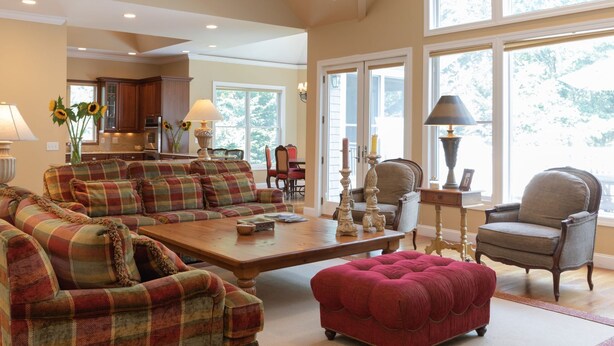 A spacious living room with a large sofa set, a red ottoman, and a wooden coffee table. There are multiple windows providing natural light and a view of the outdoors. - Bridgehampton gem in private community- 1 mile from the village (Bridgehampton)