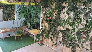 Une pièce dotée d’un sol recouvert d’un gazon artificiel vert, d’un banc en bois avec des coussins, d’une petite table et d’un mur décoré de diverses plantes et d’un motif représentant un arbre.